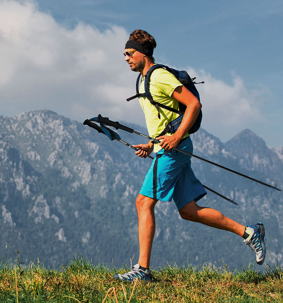 A man hiking on a mountain trail, wearing a bright yellow shirt and blue shorts. He carries trekking poles and has a backpack, with a mountainous landscape and cloudy sky in the background.