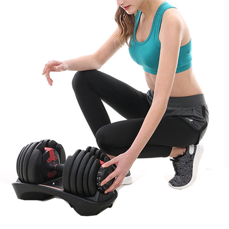 A woman in workout clothes kneels on one knee while adjusting the weight on a black adjustable dumbbell placed on its stand against a white background.
